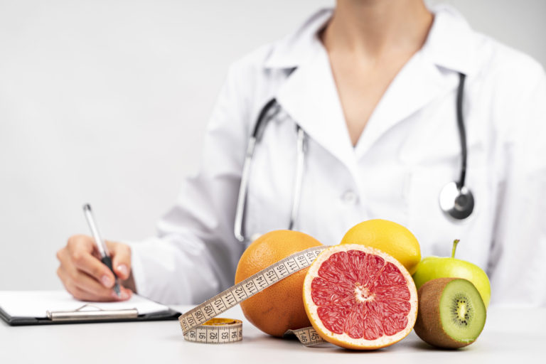 porção de frutas a frente, no fundo pessoa de jaleco preenchendo uma receita nutricional